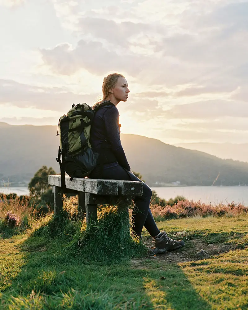 Woman with backpack sitting on a bench outdoors with mountains and water at sunset.