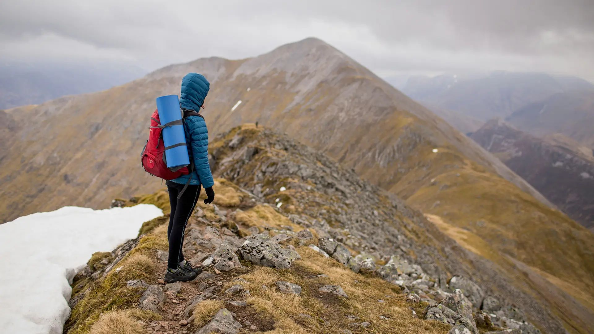 Hiker in blue jacket and red backpack standing on rocky mountain ridge near snow patch.