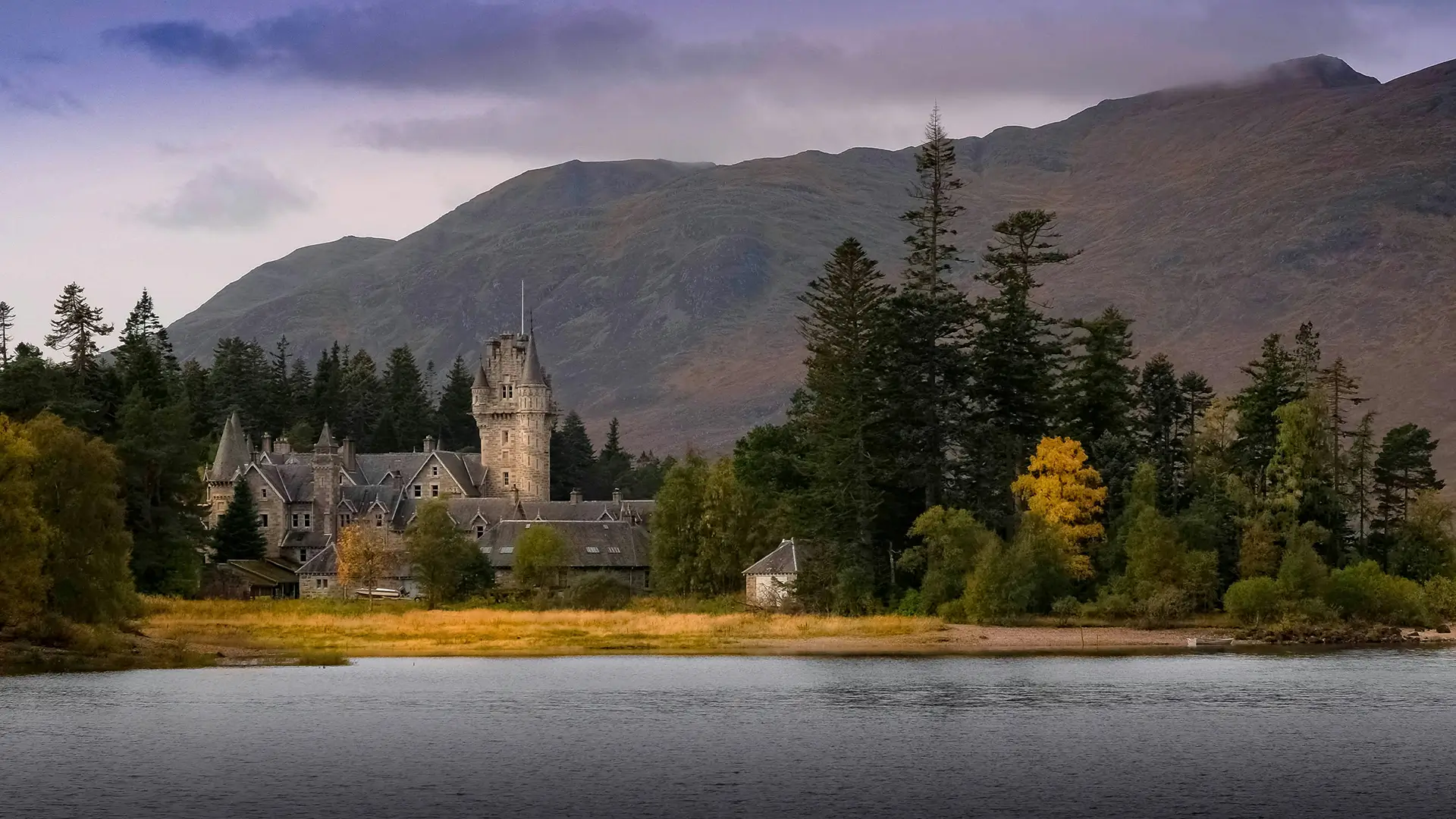 Stone castle near a forested shoreline with mountains under a cloudy sky.