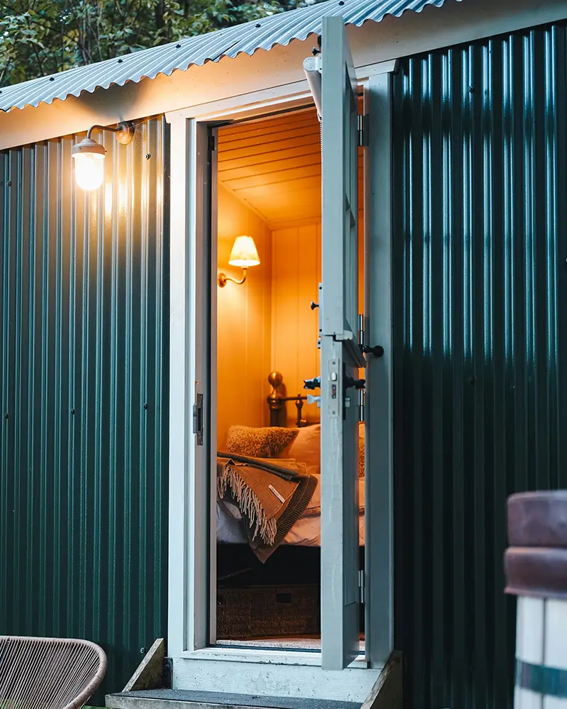 Open door to cozy cabin bedroom with lit wall lamp and a blanket on bed inside.