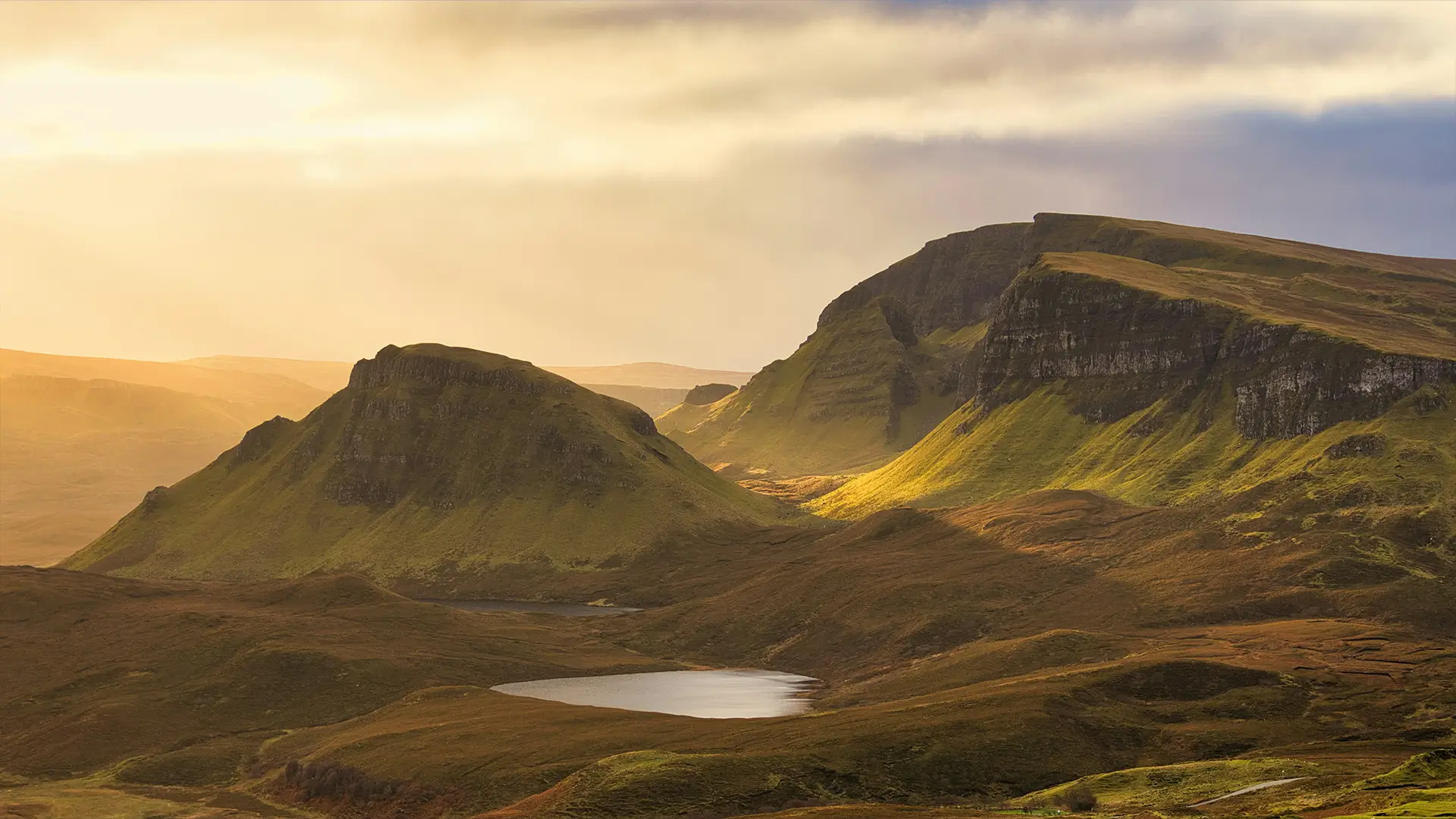 Sunlit rolling hills and cliffs with a small lake in a tranquil, foggy landscape.