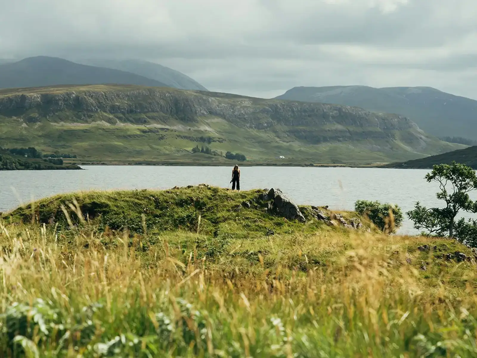 Person stands on grassy hill by a lake with mountains and cloudy sky in the background.
