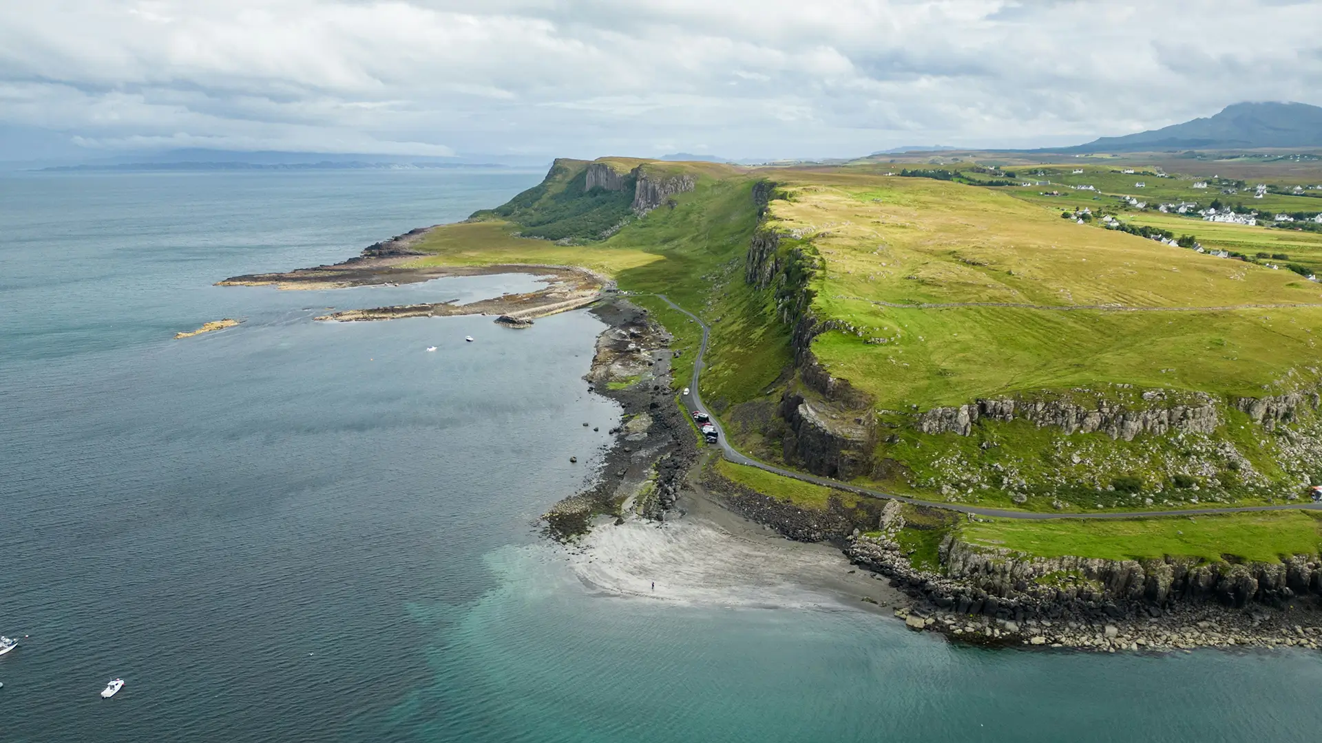 A coastal cliff with green grass and rocky shoreline beside calm blue sea under cloudy sky.