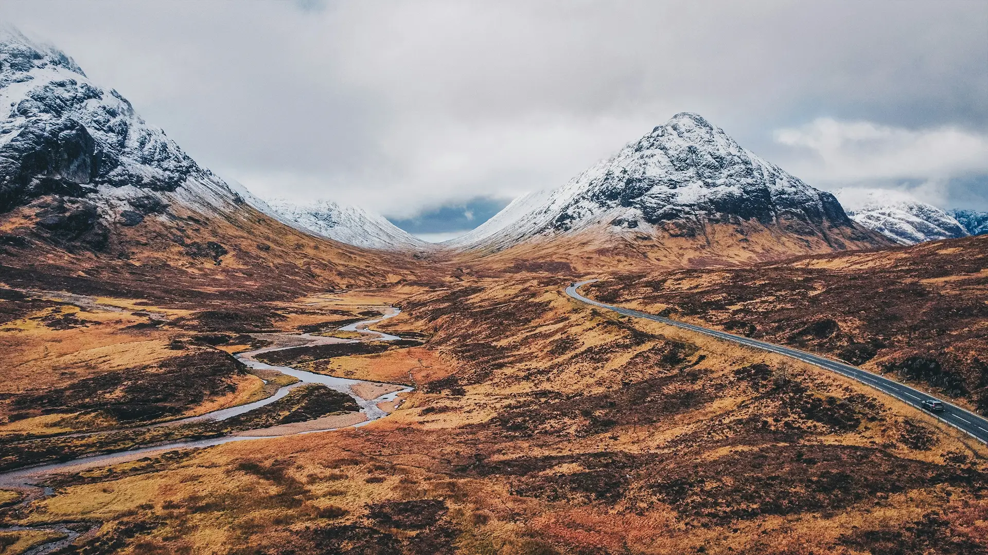 Empty road leading through golden hills with mountains and cloudy sky in the background.