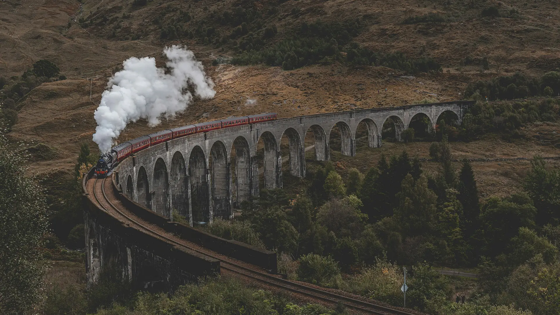 Steam train crossing a tall, curved stone viaduct in a hilly rural landscape.