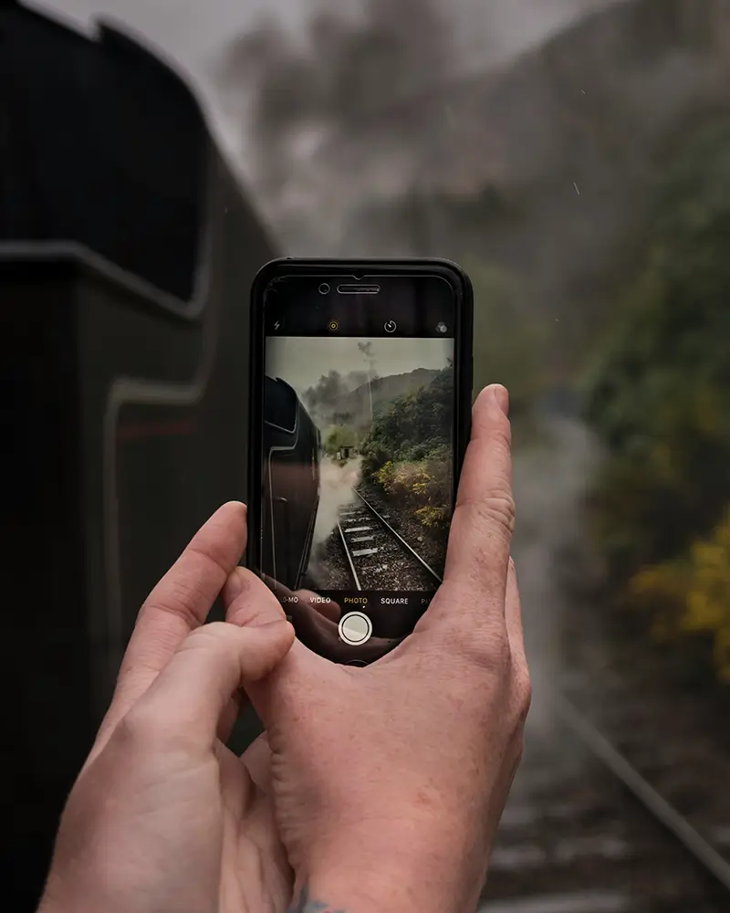 Hands holding phone capturing photo of a steam train moving on tracks through green landscape.