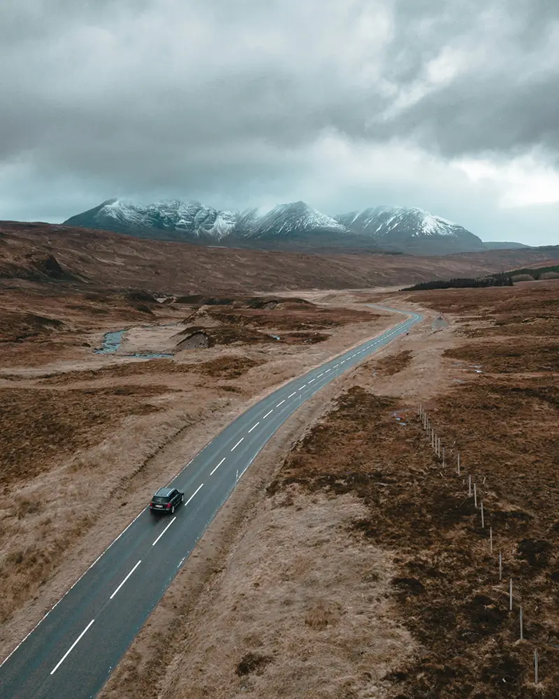 Single black car driving on winding road through barren landscape with snowy mountains under cloudy sky.