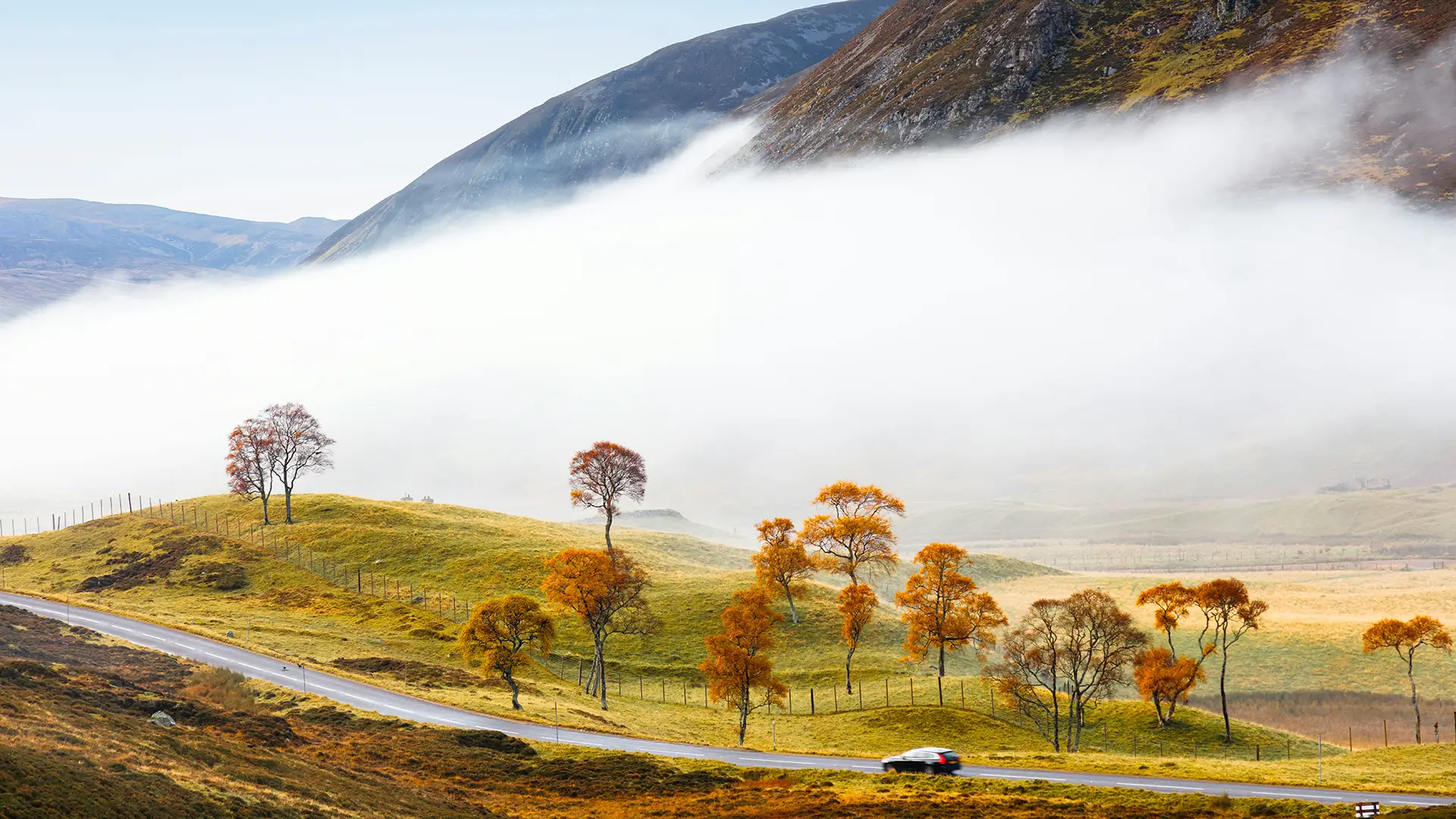 Car driving on a winding road through autumn trees and grassy hills with fog and mountains in background.