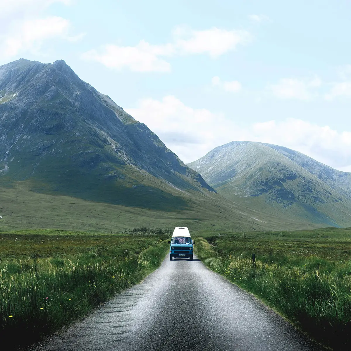 Van driving on a narrow road surrounded by green fields and mountains under a blue sky.