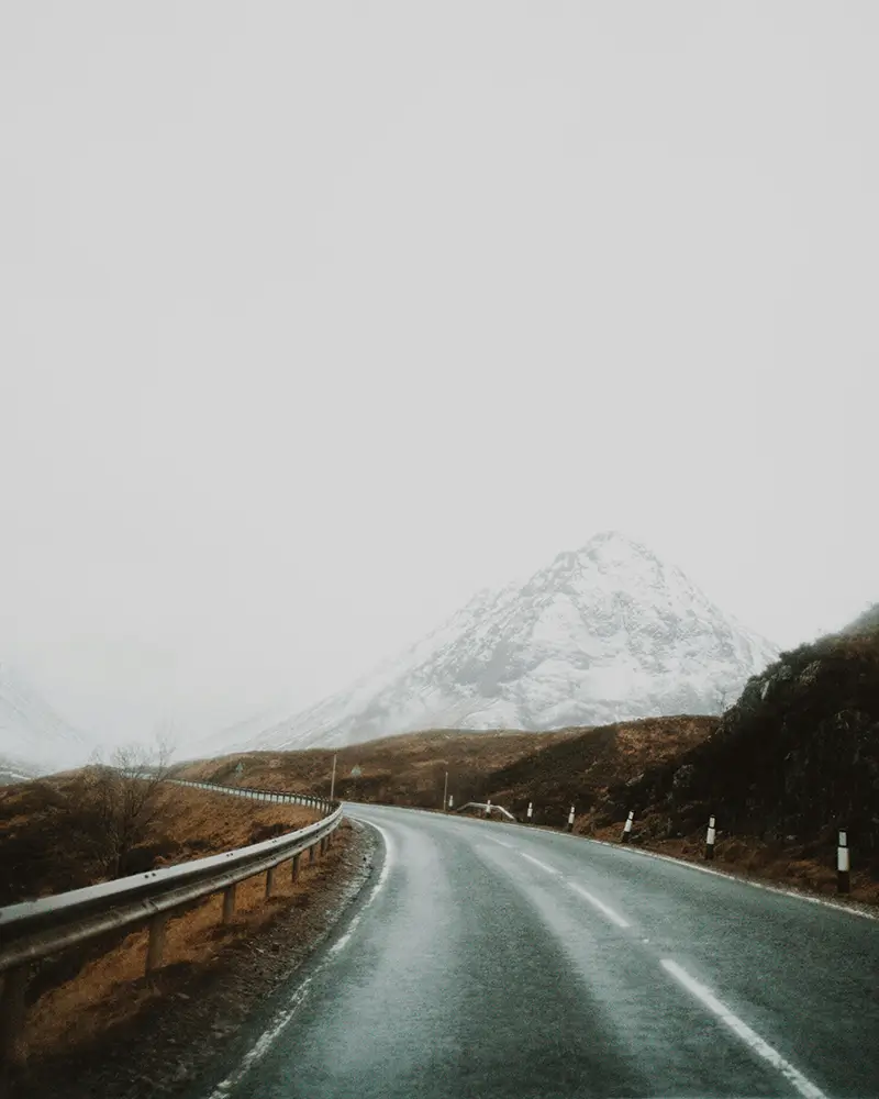 Winding road with guardrails passes through brown hills toward snowy mountain under gray sky.