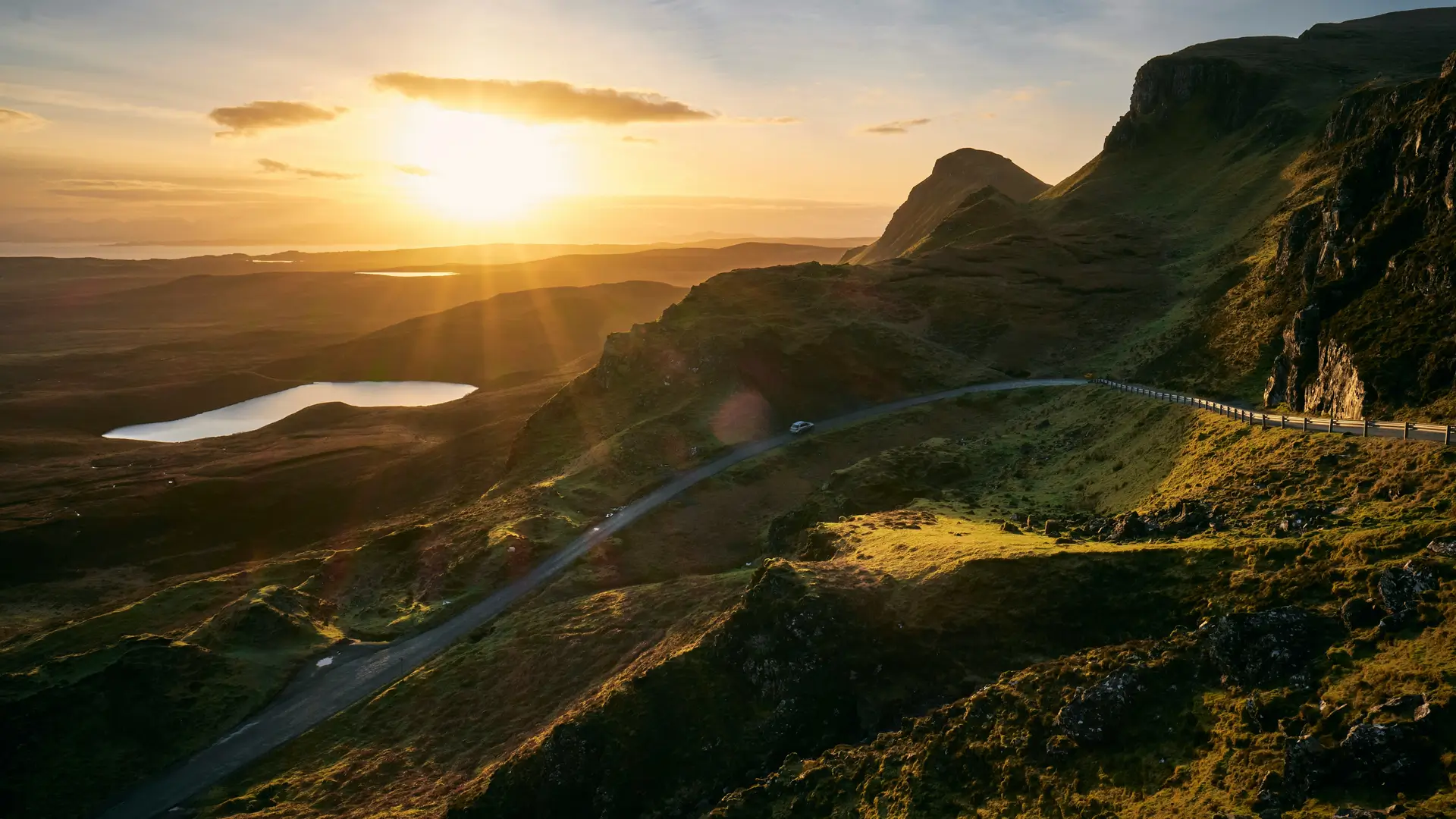 Winding road through green hills with sunset and a lake in the distance.