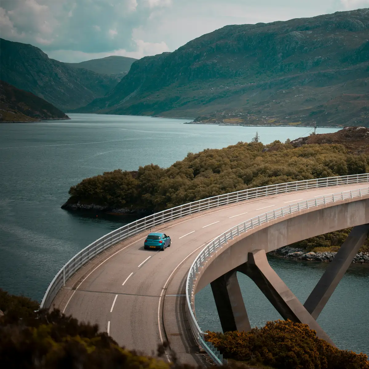 Blue car driving on curved bridge over water with mountains and trees in the background.