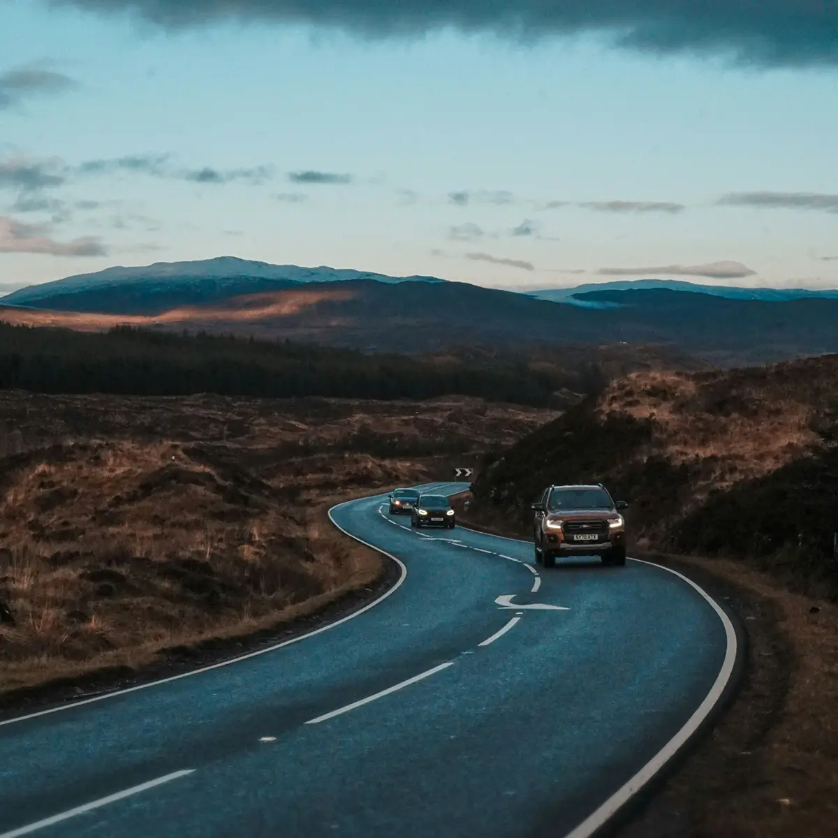 Curving road in a hilly landscape with three cars driving toward the camera under a cloudy sky.