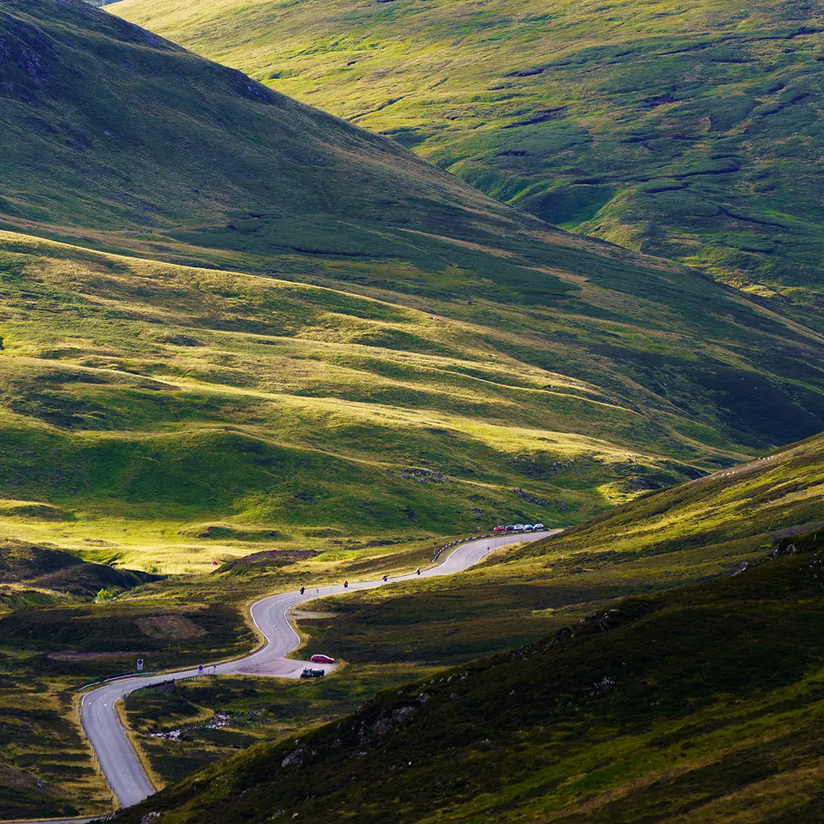 Winding road through green hills in Scotland with parked cars and motorcycles.