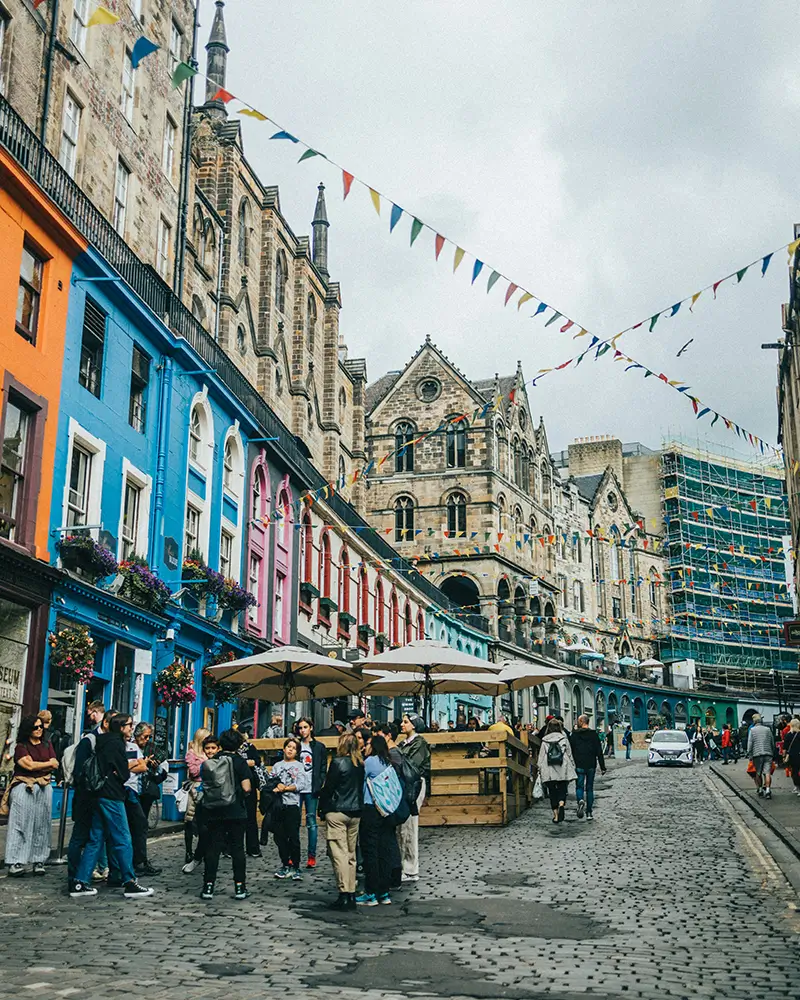 Crowd on cobblestone street with colorful buildings and historic stone architecture in the background.