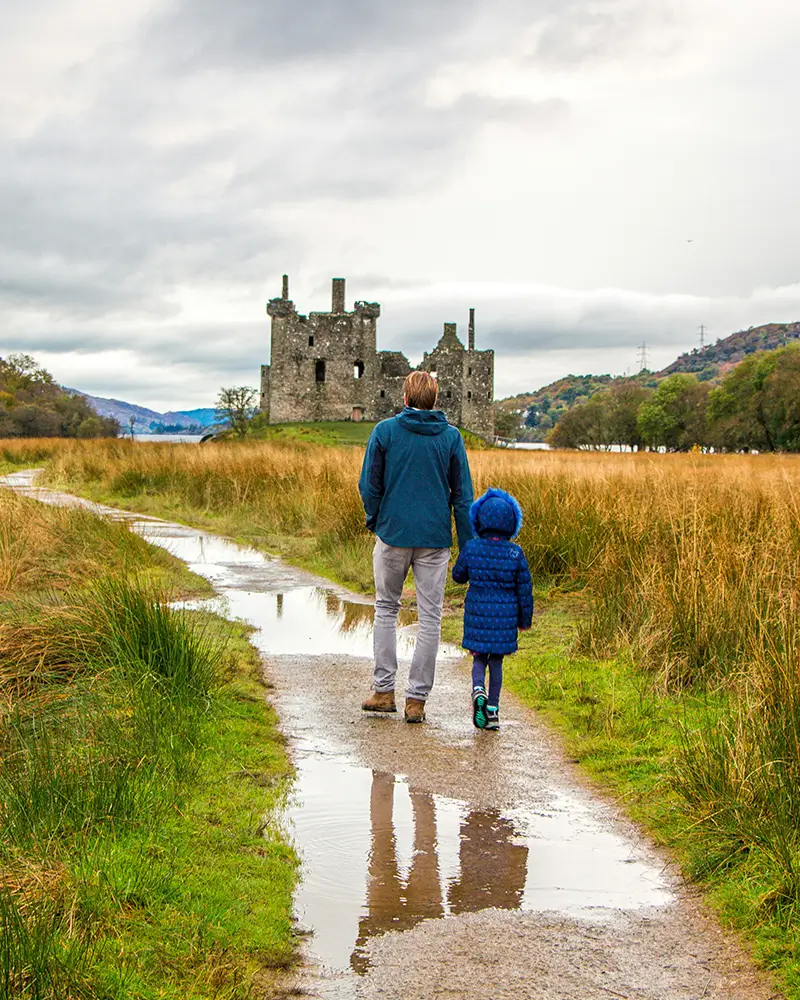 Adult and child in blue jackets walking on a wet path toward an old stone castle in a grassy field.