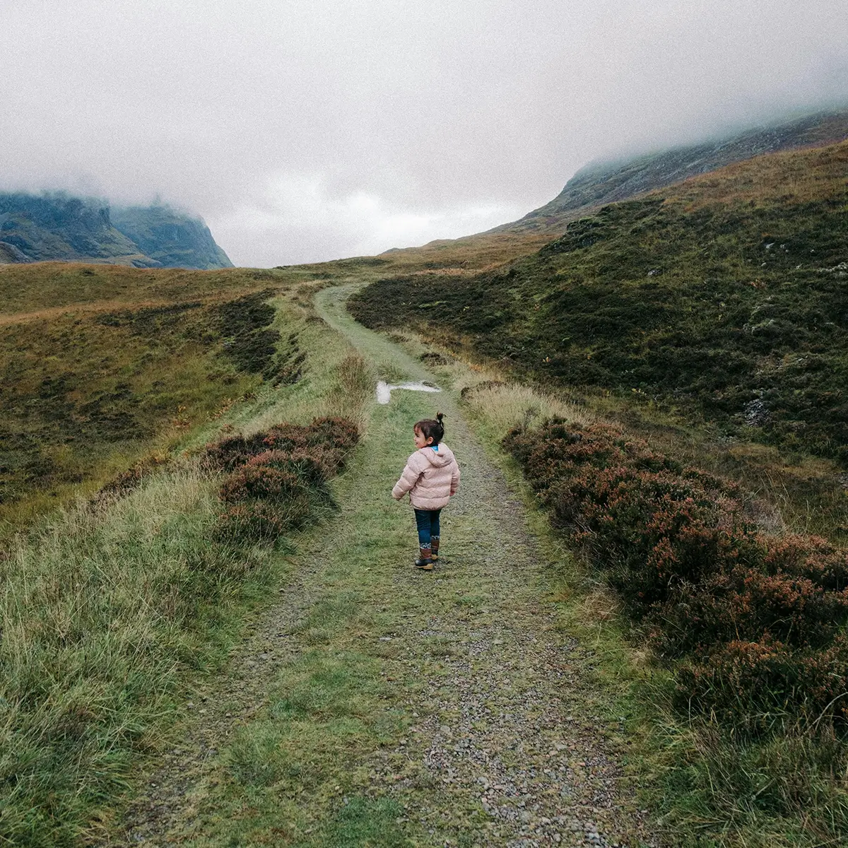 Child in a pink jacket and boots walking on a green path in misty hills.