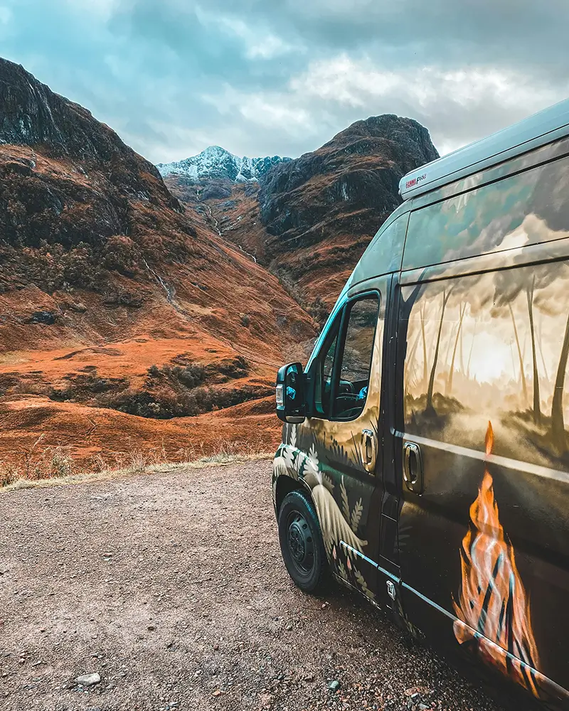 Van with nature and fire artwork parked on gravel near mountainous landscape with cloudy sky.