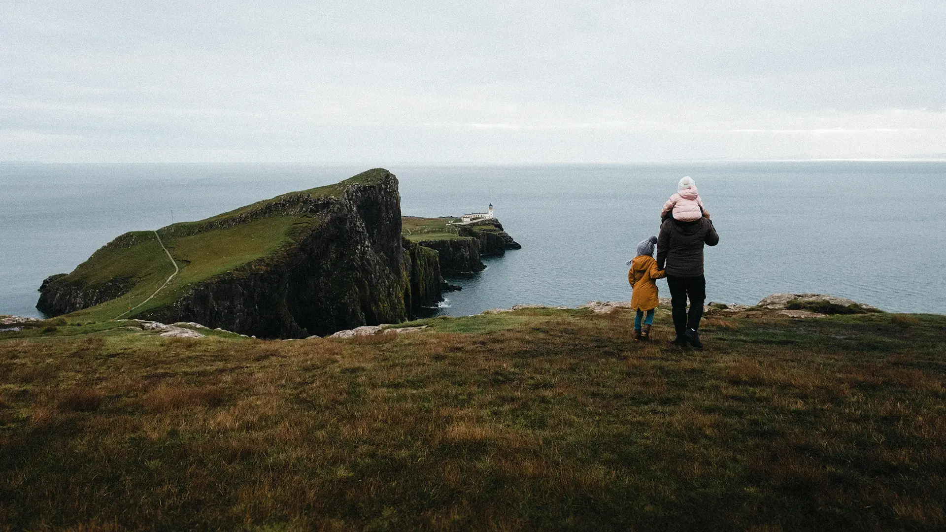 Two children in winter hats and jackets sitting on grass by cliffs overlooking sea and distant lighthouse.