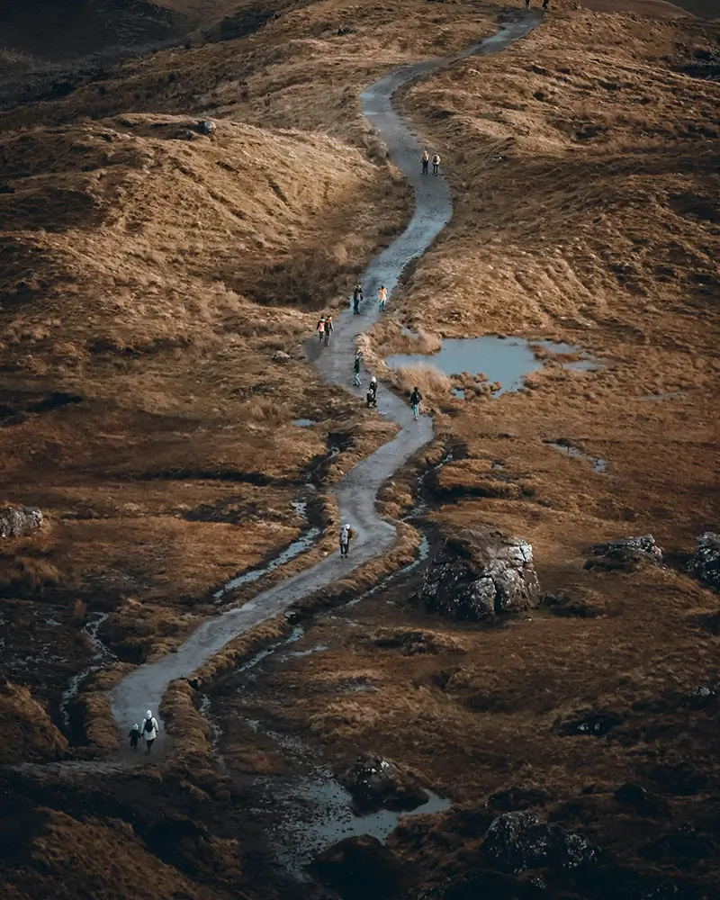A winding narrow path with people walking, surrounded by brown grassy hills and rocky terrain.