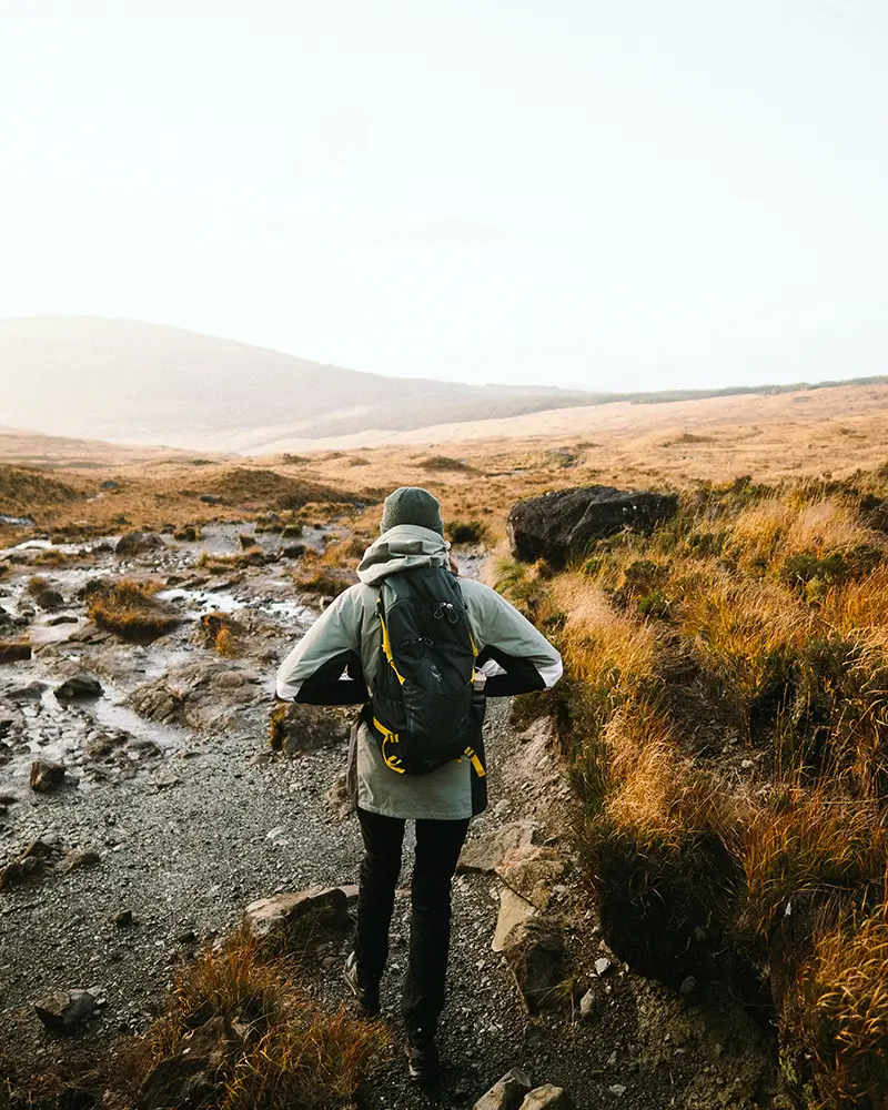 Person hiking on rocky trail through golden grassy terrain with hills in the distance.