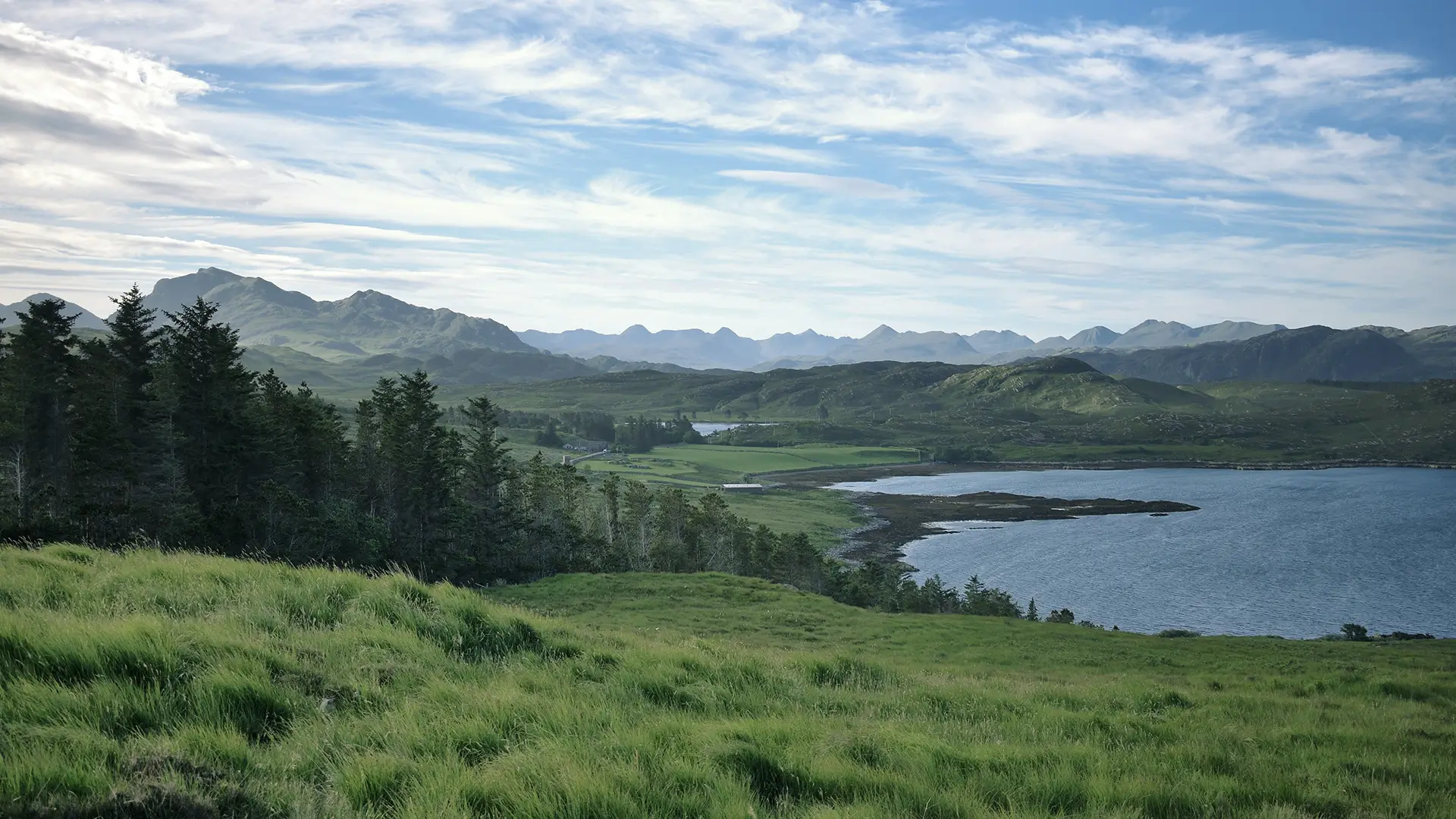 Scenic Scottish landscape with green hills, forest, coastal water, and distant mountain range under a cloudy sky.