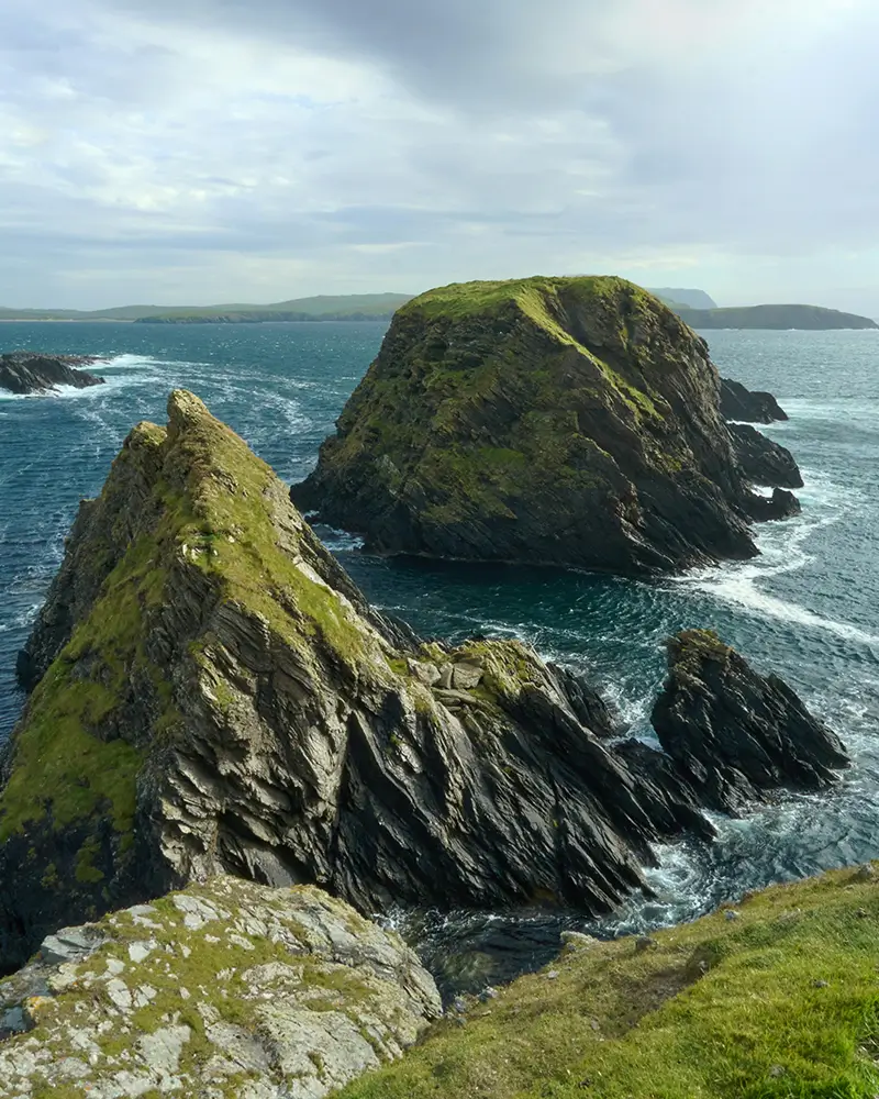 Rocky grassy islands in blue ocean under a cloudy sky with distant land visible on horizon.
