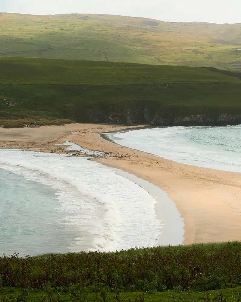 Curved sandy beach with waves and green hills in the background under cloudy sky.