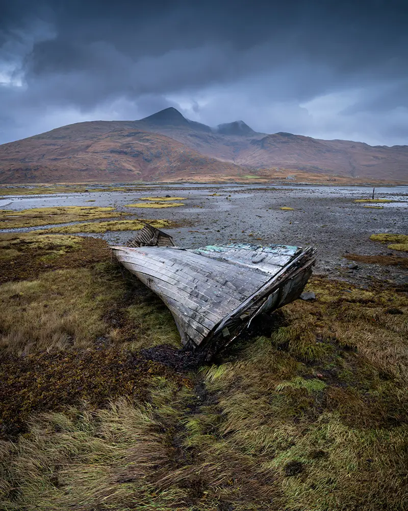Weathered, overturned wooden boat lying on grassy shore under cloudy sky by distant hills.