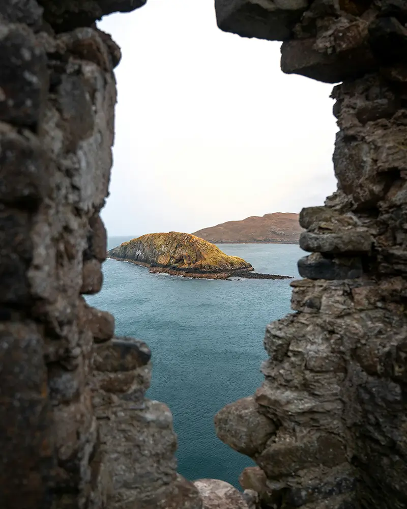 View of a small island and water through a jagged stone window of an old castle.
