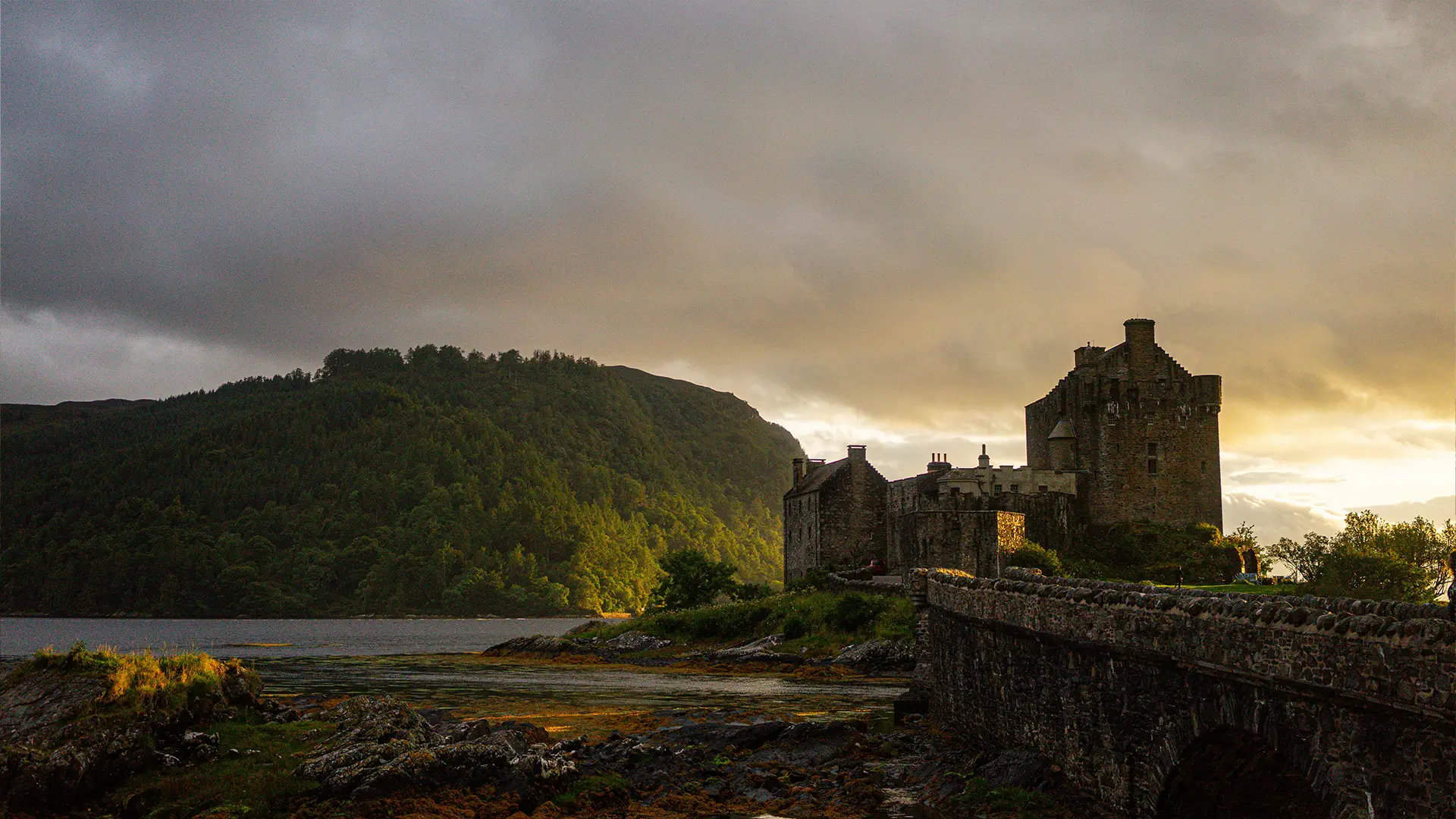 Historic castle on an island surrounded by water, autumn trees, and distant hills under a cloudy sky.