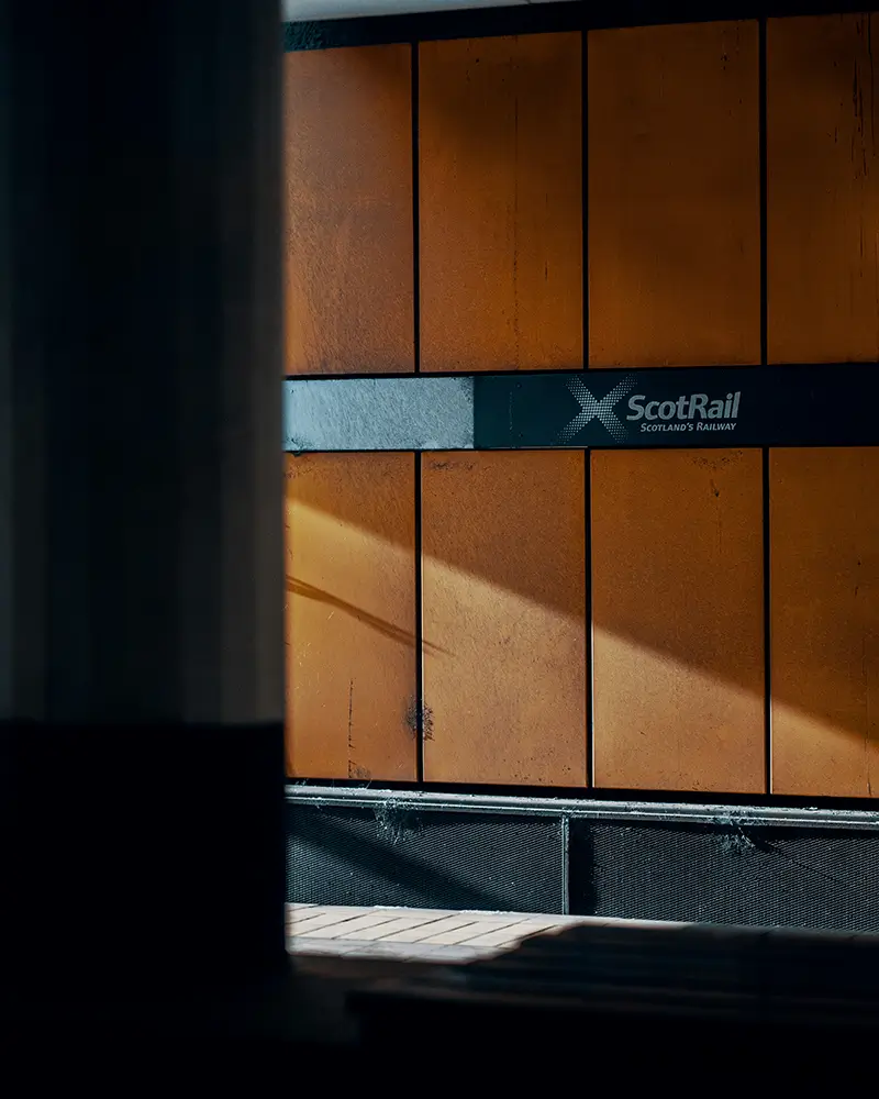 Sunlight casting shadows on orange panels with ScotRail logo and tagline on a station wall.