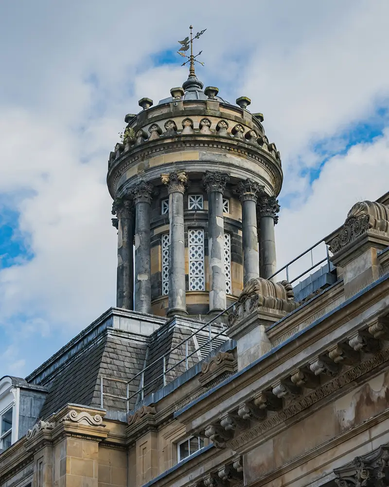 Ornate stone tower with columns and weather vane on top of historic building under blue sky.