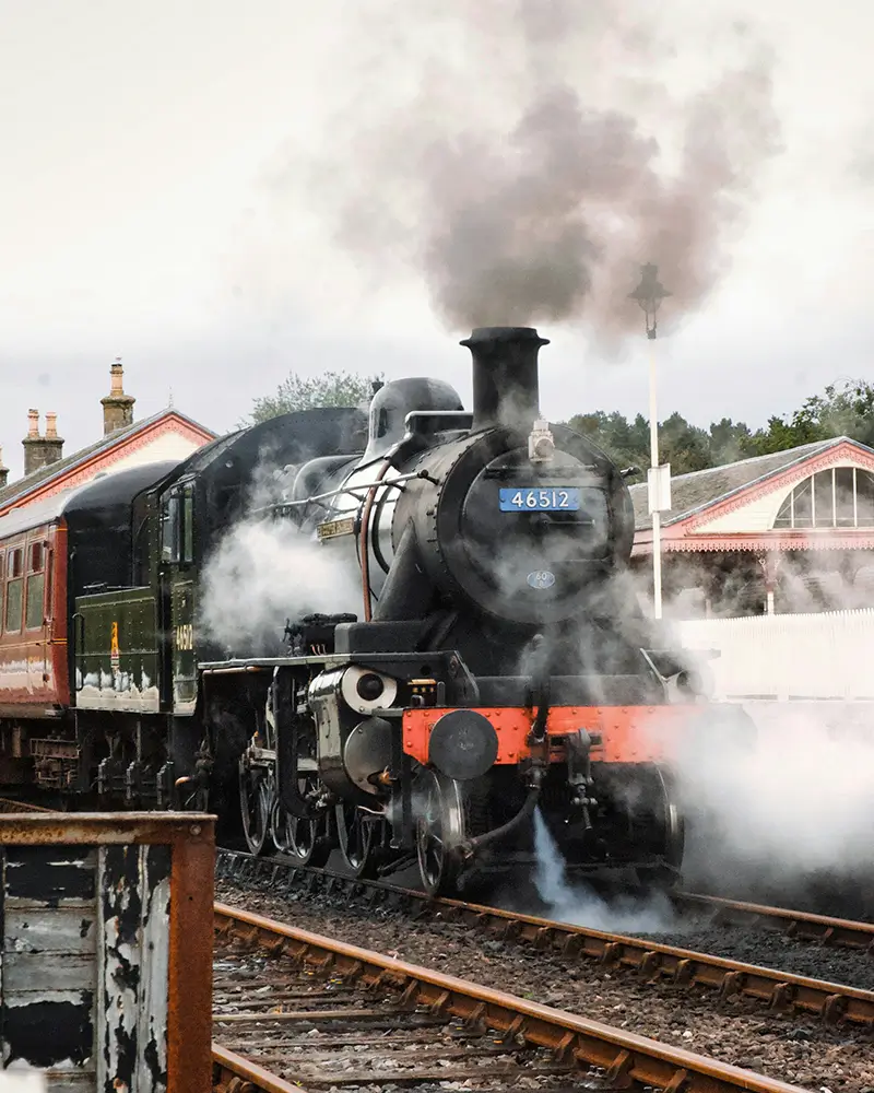 Black vintage steam train emitting smoke at a railway station platform with red-trimmed roof buildings.