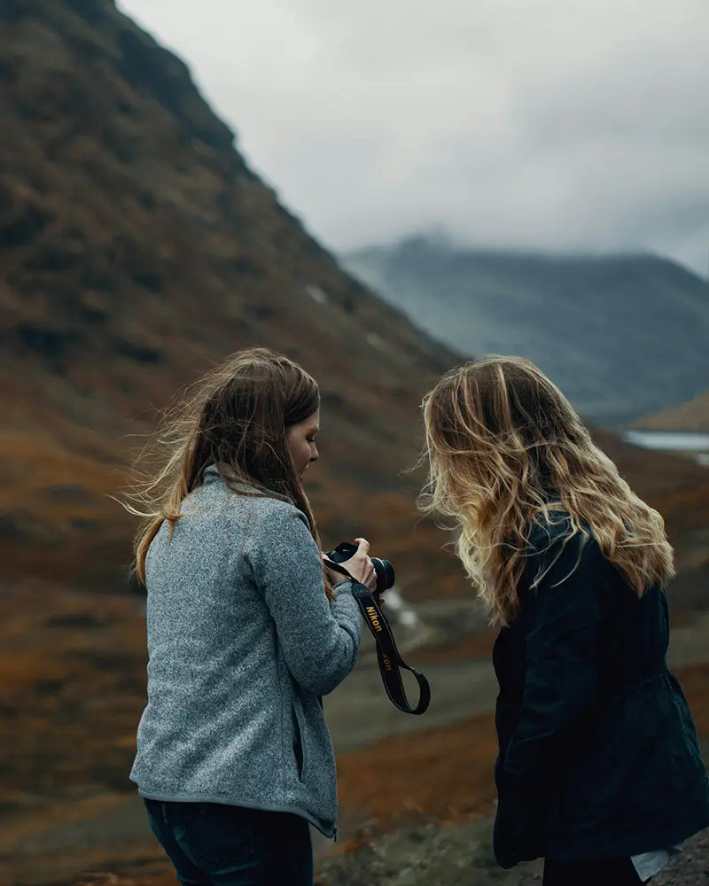 Two women outdoors with a camera, mountains and cloudy sky in the background.