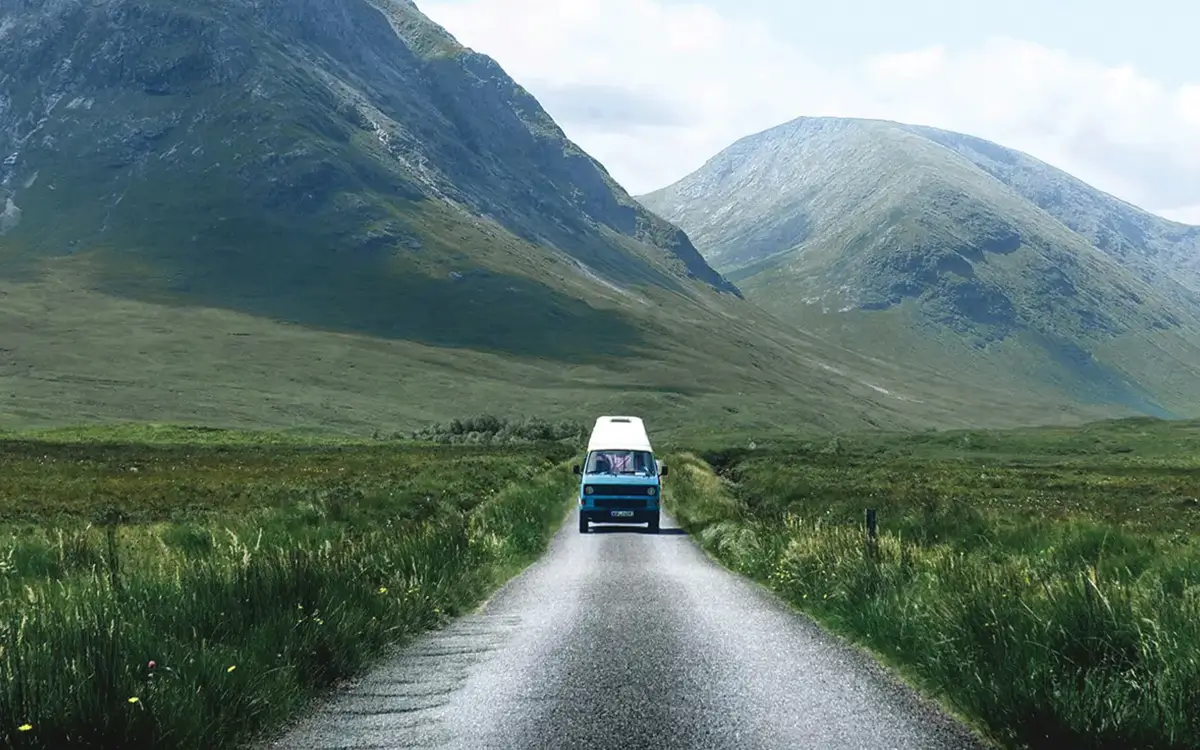 Blue camper van driving on a narrow road through green fields with large mountains in the background.