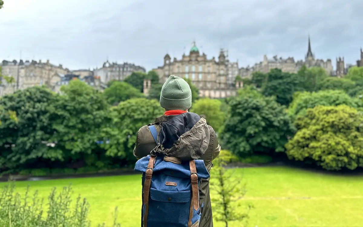 Person wearing a green beanie and blue backpack looks at a cityscape beyond green trees and grass.