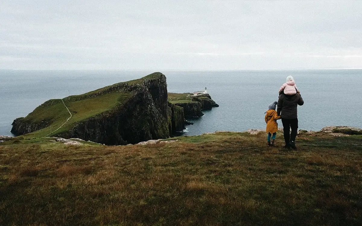 Adult with two children overlooking cliffs and lighthouse by the sea on cloudy day.