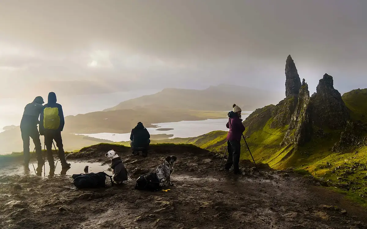 Hikers with dogs and cameras on a muddy cliff overlooking rocky peaks and foggy lake in Isle of Skye.
