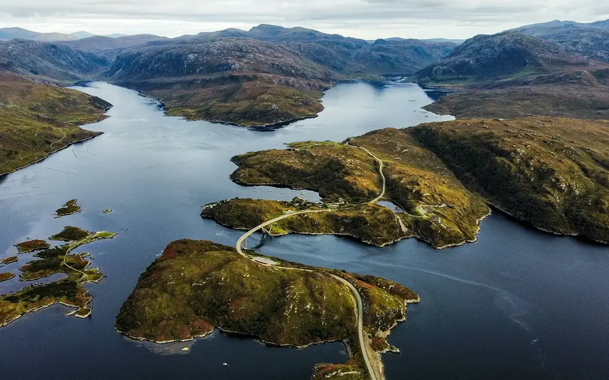 Aerial view of a winding bridge over blue water connecting green hills and mountainous coastline.