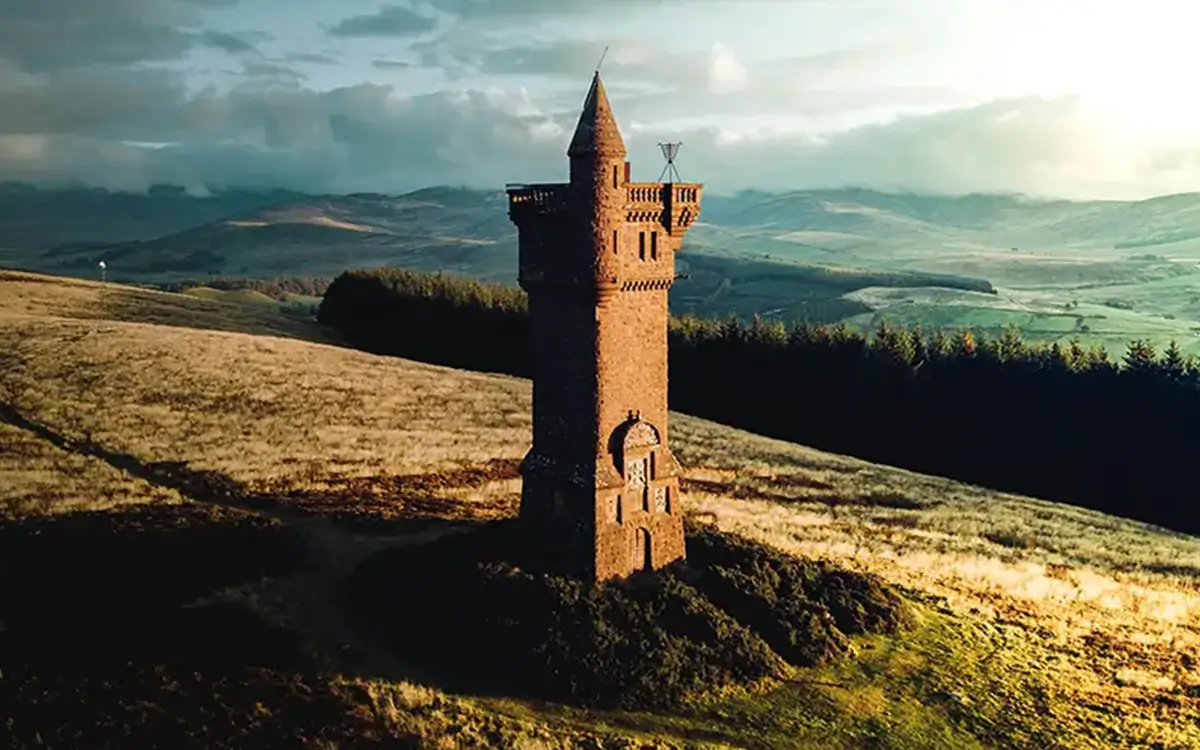 Stone tower with battlements on grassy hillside under a cloudy sky with distant hills.
