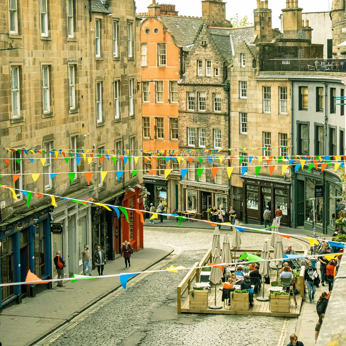 Cobblestone street with outdoor café and people walking under colorful pennant flags in historic buildings.