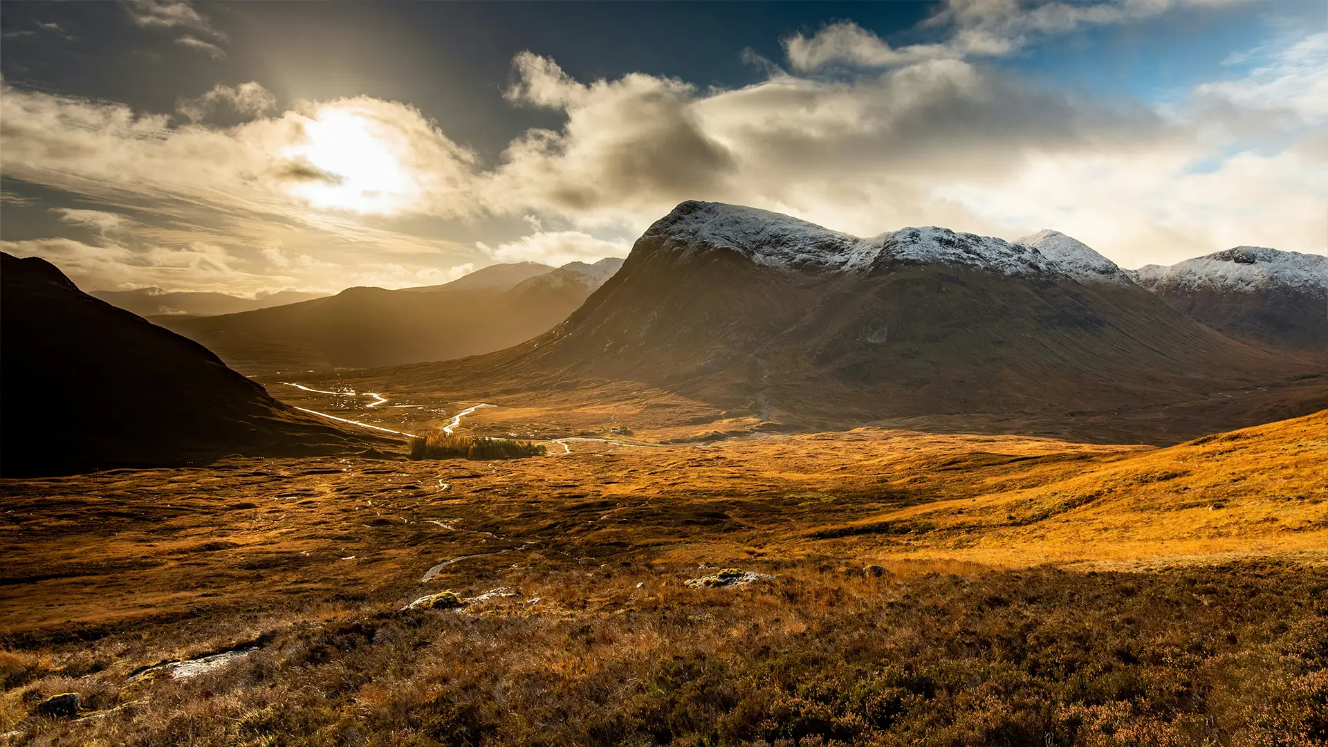Sunset over a golden valley with a winding river and snow-capped mountains under a cloudy sky.