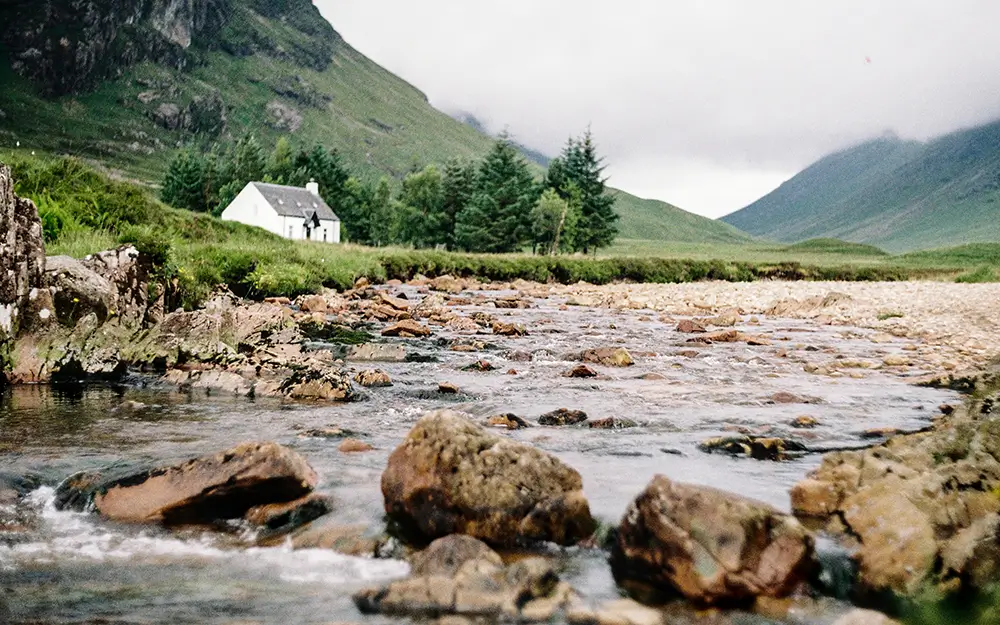 Stream with rocks flowing through a green valley with a white house and hills in the background.