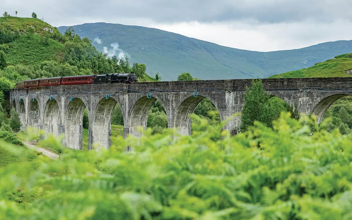 Steam train crossing a stone arched viaduct in lush green hills under a cloudy sky.