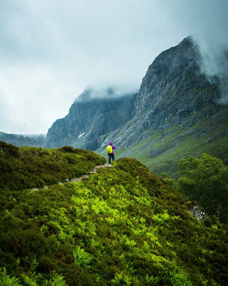 Hiker with bright backpack on green trail below misty, rocky mountain peaks.