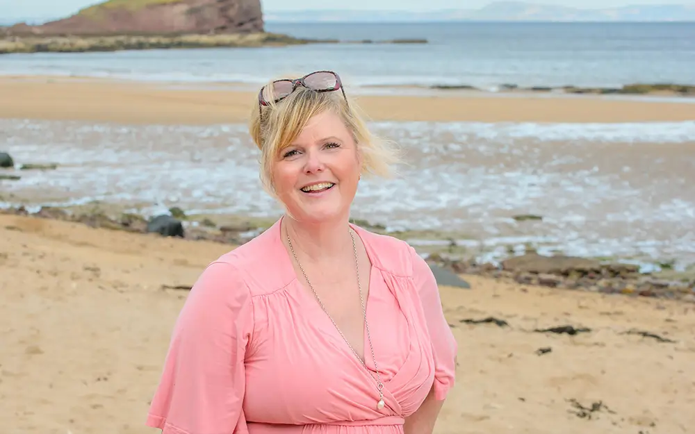 Smiling woman in pink dress with glasses on her head standing on a sandy beach near the water.