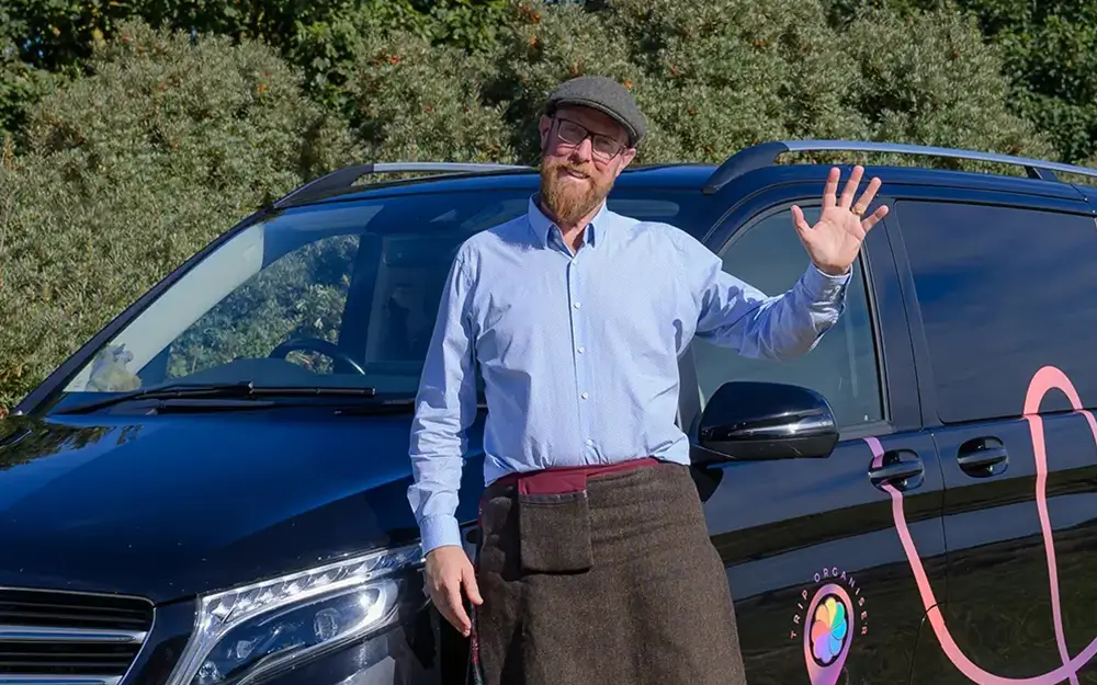 Smiling man in a cap and kilt waving while standing next to a black vehicle with branding.