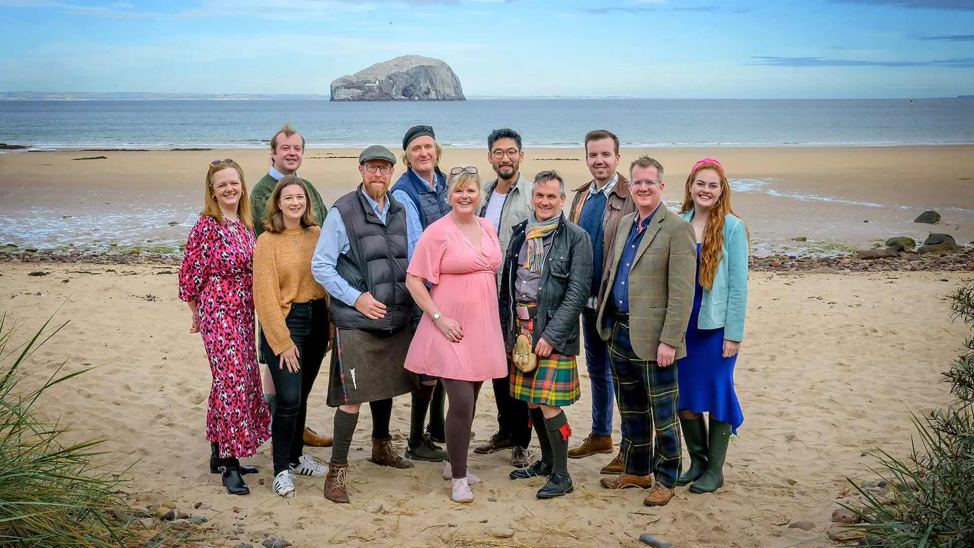 Group of eleven people standing on a sandy beach with a large rock island in the sea behind them.