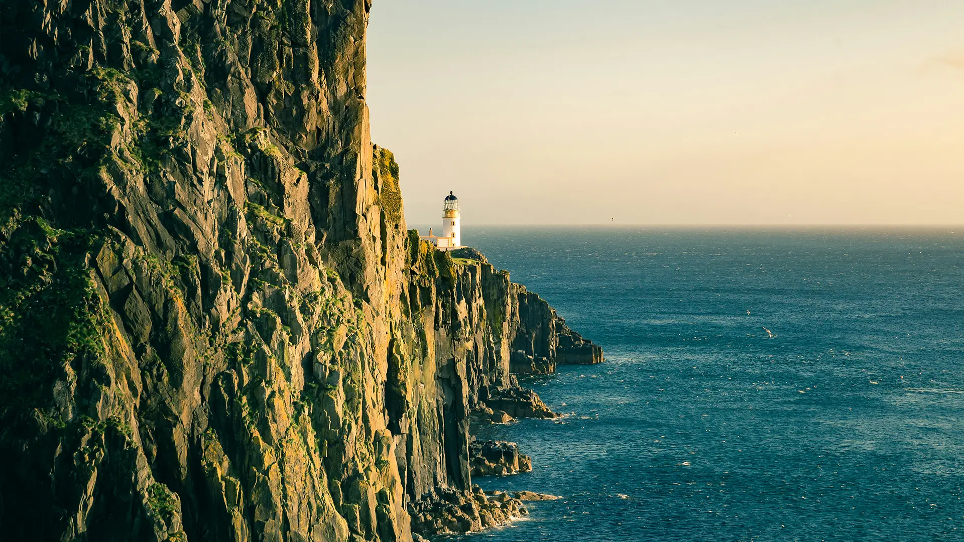 White lighthouse on the edge of a rocky cliff overlooking a calm blue sea at sunset.